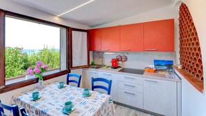 Eine kleine Küche mit weißen und orangefarbenen Schränken, einem Esstisch mit blauen Stühlen und Blick ins Grüne durch ein Fenster.