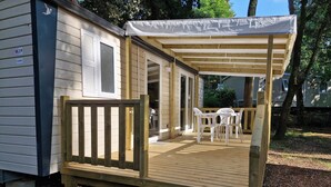 Una terraza de madera con un porche cubierto, una pequeña mesa con sillas y un escalón que lleva a una puerta.