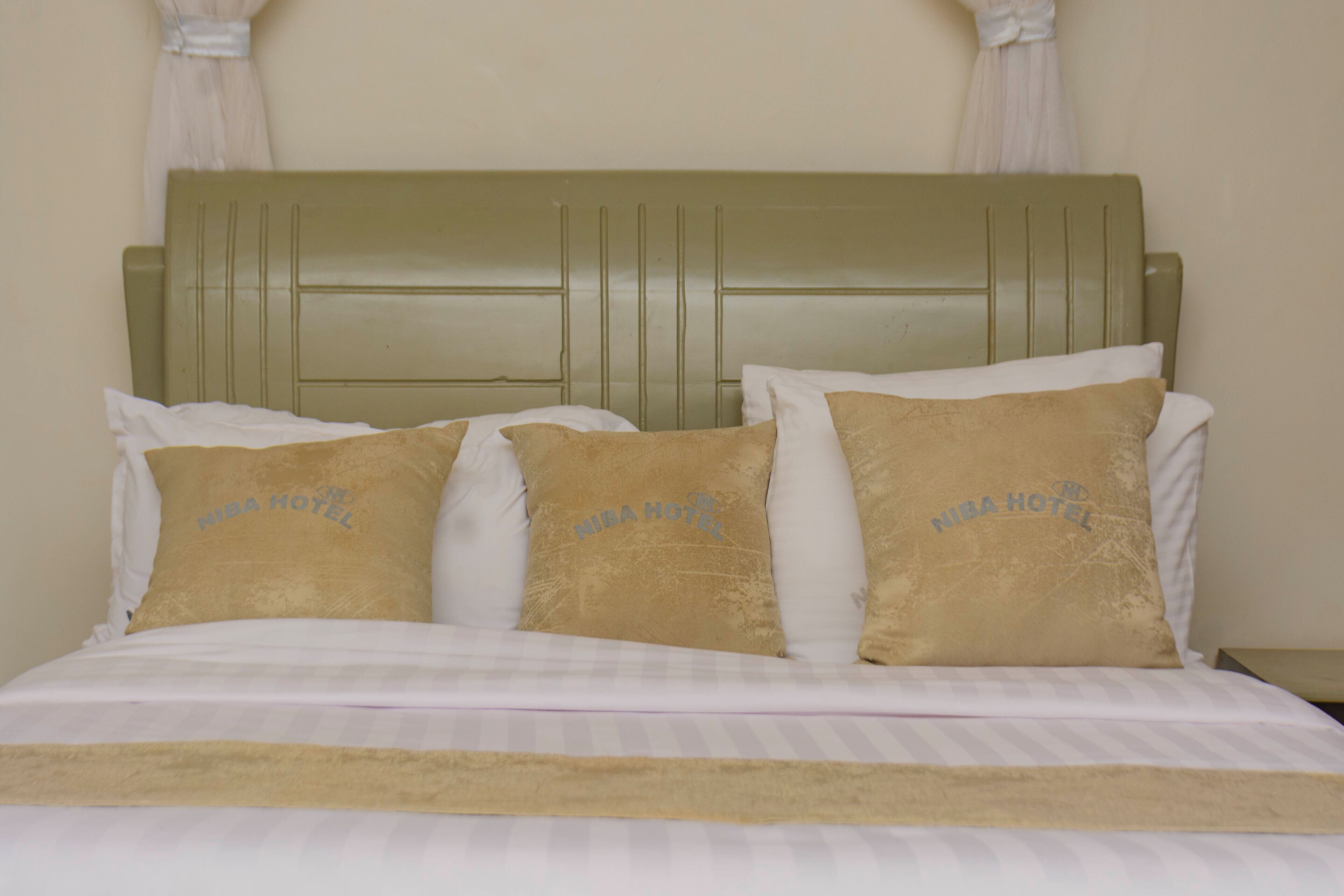 A bed with a green headboard, white bedding, and three beige pillows with "NIBA HOTEL" text.