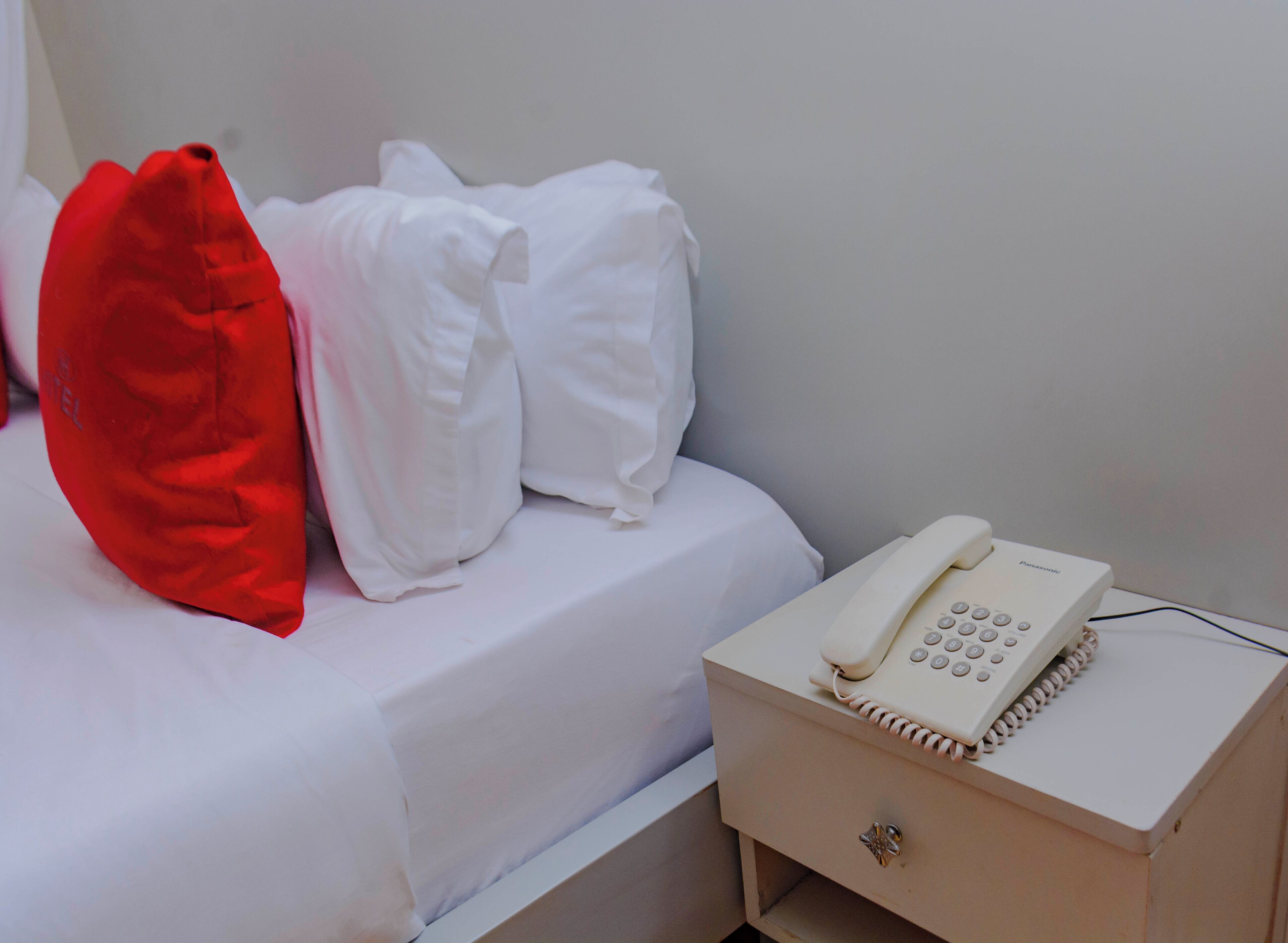 Uma cama com roupa de cama branca e um travesseiro vermelho, um telefone branco na mesa de cabeceira e uma pequena mesa de cabeceira de madeira.