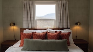 A bedroom with a large window, patterned wallpaper, a bed with multiple pillows, two bedside lamps, and a nightstand.