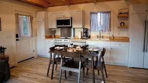 A compact kitchen with white cabinets, a microwave, a stove, and a refrigerator. There is a dining table with chairs and a window with checkered curtains.
