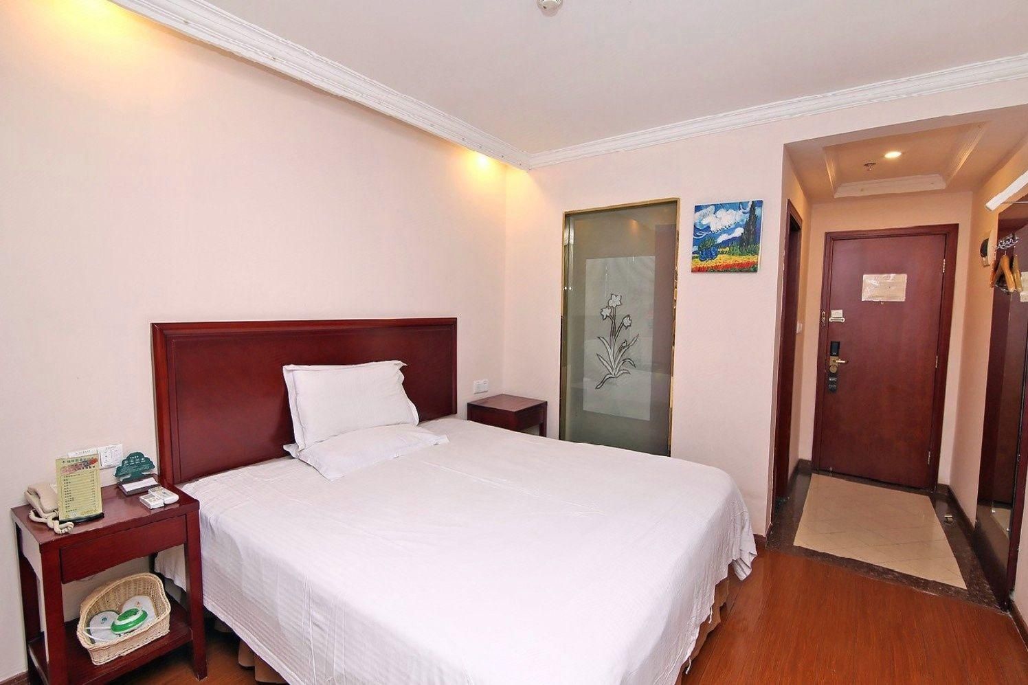 Une chambre d’hôtel avec un grand lit, une table de chevet, un téléphone fixé au mur, une porte avec un panneau en verre et un petit tableau.