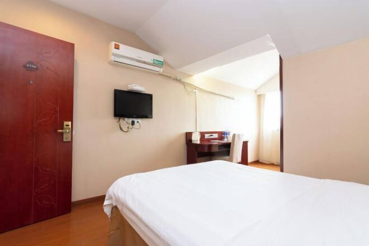 Ein Hotelzimmer mit einem Bett, einem an der Wand befestigten Fernseher, einem Schreibtisch mit Stuhl und einer Klimaanlage.