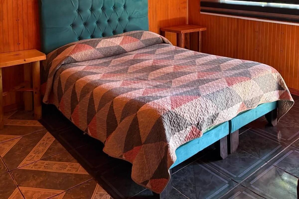 Une pièce avec un plafond en bois, une tête de lit capitonnée de couleur turquoise, une courtepointe à motifs et une fenêtre avec des stores.