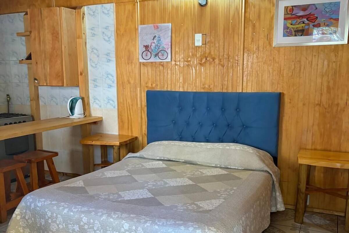Une chambre avec une tête de lit bleue, un lit recouvert d’une courtepointe à motifs floraux, des murs en bois, un petit bureau et une chaise.