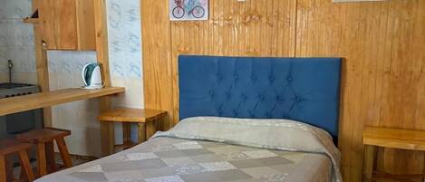 Un dormitorio con cabecera azul, cama con cubrecama floral, paredes de madera, un escritorio pequeño y una silla.