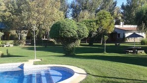 A swimming pool with a geometric tile pattern, surrounded by a well-maintained lawn and a small tree. - casa rural la badia (Lucillos)