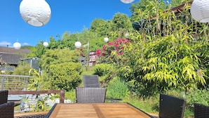 An outdoor dining area with wicker furniture and a wooden deck, surrounded by lush greenery and flowering plants. - Vacation apartment with heart, for up to 3 people & dog in the heart of nature (Allendorf/Eder)
