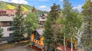 A mountain village with traditional architecture, including a yellow building with a balcony and flower boxes, surrounded by trees and set against a backdrop of hills. - Lift House Lodge, Downtown Vail Lionshead Village Studio Condo (Vail)