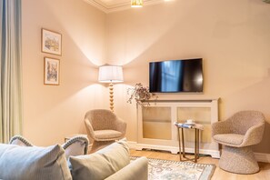 Een knus woongedeelte met een bank, twee fauteuils, een televisie boven een open haard, en een bijzettafeltje met een lamp.