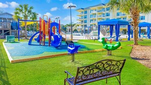 Un parque infantil con toboganes y columpios, un banco y un edificio con balcones al fondo.