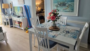 Comedor con mesa blanca dispuesta para cuatro, un jarrón de flores y un cuadro enmarcado sobre la mesa.