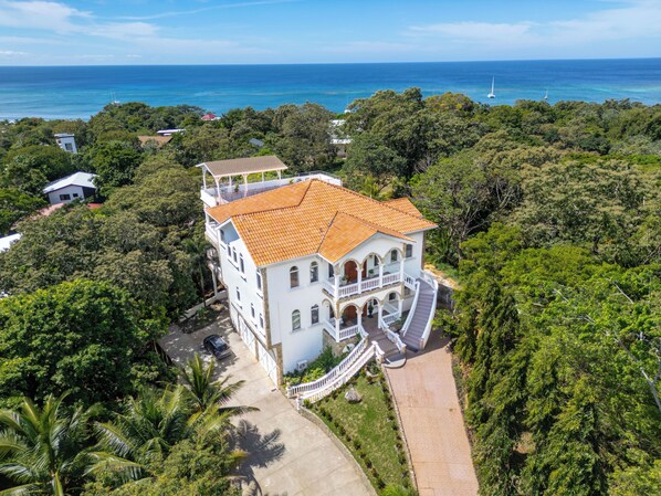 A large white building with a red-tiled roof, surrounded by lush greenery and a view of the ocean.