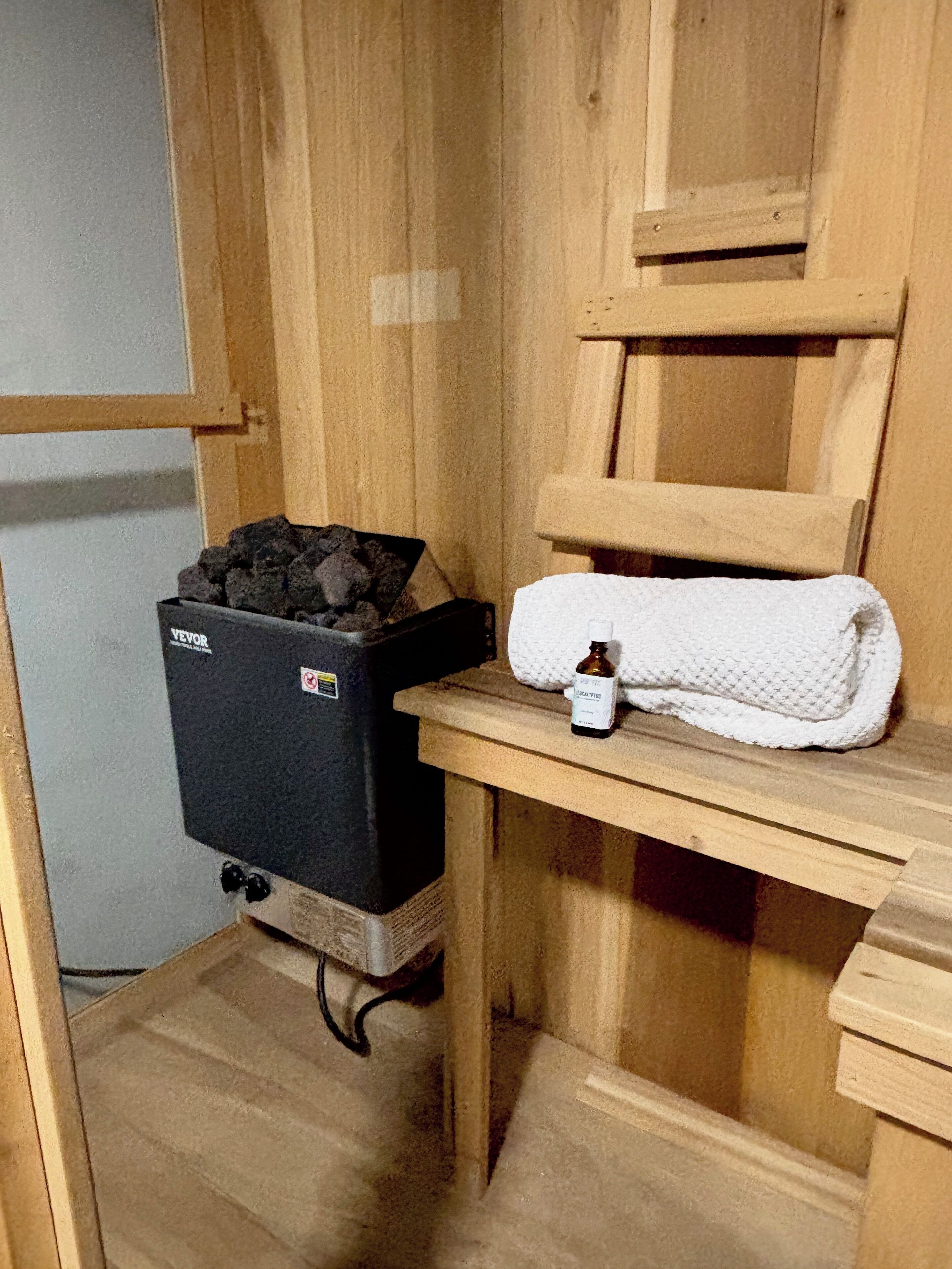 A bathroom with a black trash bin, a white towel, and a small bottle on a wooden shelf.