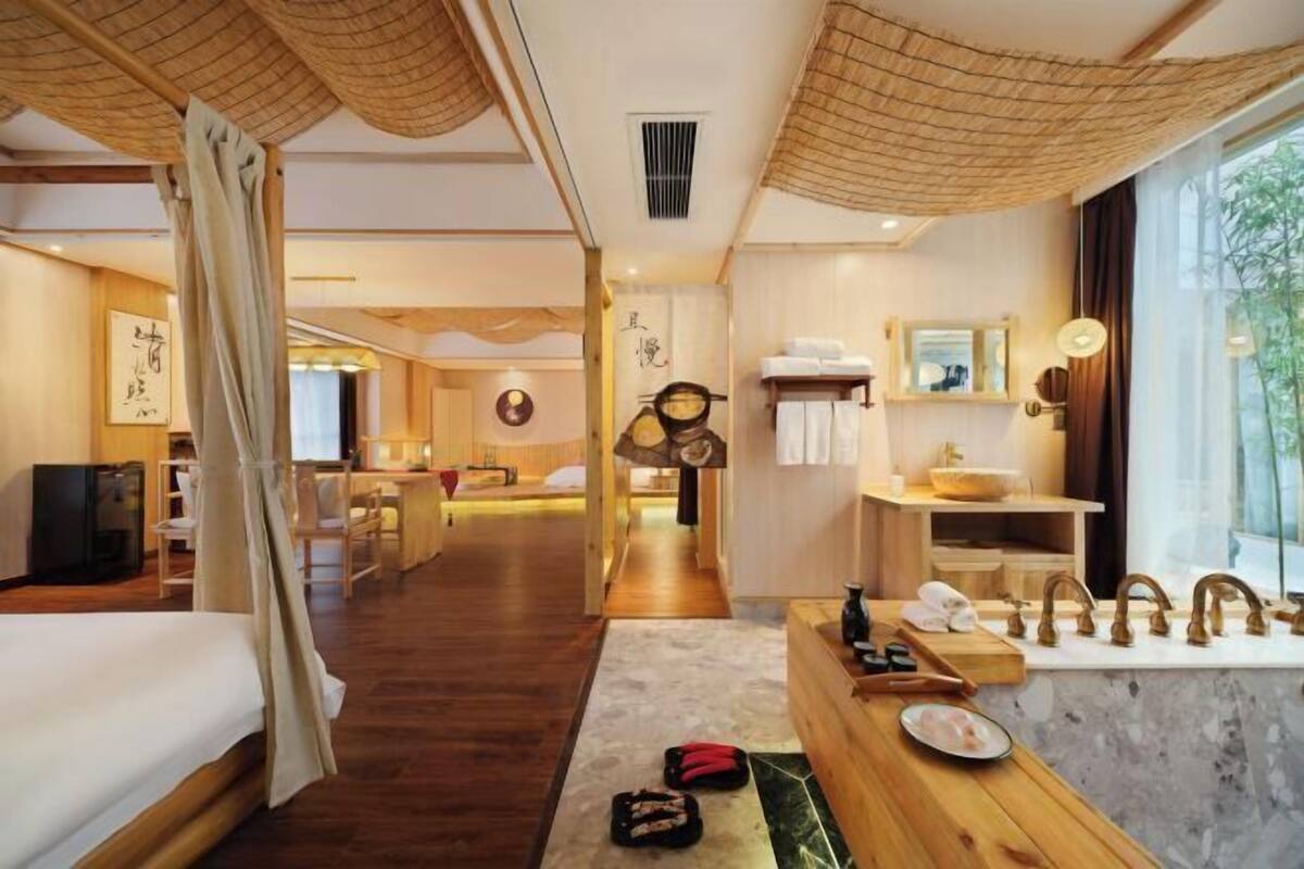 Ein Hotelzimmer mit Bett, einem Holztisch, einem Badezimmer mit Waschbecken und Spiegel sowie Blick ins Freie.