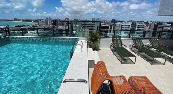 Una piscina en la azotea con agua azul cristalina, rodeada de sillones de descanso y vistas a la ciudad.