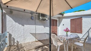 Un patio con mesa y sillas, una sombrilla beige y una pared blanca con contraventanas.