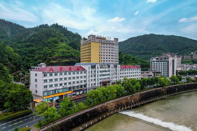 A multi-story hotel with a red roof, situated beside a river with a dam, surrounded by greenery and hills.
