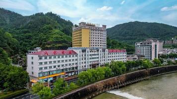 A multi-story hotel with a red roof, situated beside a river with a dam, surrounded by greenery and hills.