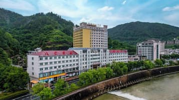 A multi-story hotel with a river in the foreground, surrounded by greenery and hills.