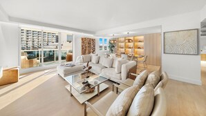 A spacious living room with a large sofa, a coffee table, and a dining area. There is a large window with a view of a building.