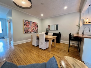 A modern living room with a wooden dining table, white chairs, a large abstract painting, a TV, and a kitchen area with a sink and countertop. - Montréal Haven (Montréal)