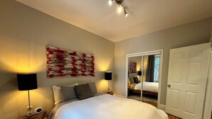 A bedroom with a large bed, two bedside lamps, a wooden nightstand, a mirror, and a door. - Montréal Haven (Montréal)