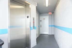 Un pasillo con un ascensor, señal de salida y una puerta con placa de número.