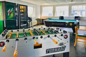 A room with a foosball table labeled 'TORNADO,' a pool table, a vending machine with a 7UP logo, and a window providing a view of the outside. - OCEAN VIEW Studio@Daytona Zzz 4 (Daytona Beach)