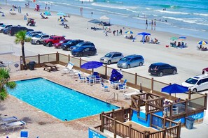 A pool area with lounge chairs and umbrellas, adjacent to a beach with parked cars and umbrellas. - OCEAN VIEW Studio@Daytona Zzz 4 (Daytona Beach)