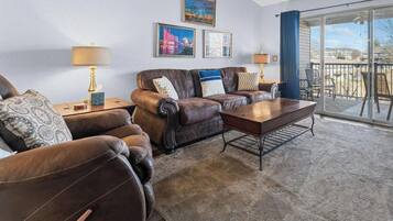 A living room with a brown leather sofa set, a wooden coffee table, and two lamps on side tables. There are framed pictures on the wall and a balcony with a view.