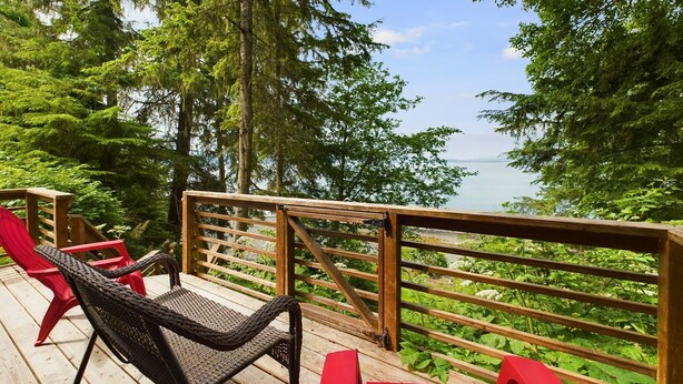A wooden deck with wicker chairs and red cushions, overlooking a forested area and a body of water. - Lena Beach Retreat - Oceanfront Luxury (Juneau)