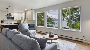A modern kitchen with white cabinets and a large island, a gray sofa, and a view of a lake through large windows. - Lena Beach Retreat - Oceanfront Luxury (Juneau)