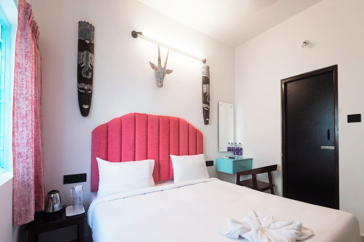 A hotel room with a large bed, a red upholstered headboard, a bedside table with a lamp, a mirror, and a door.