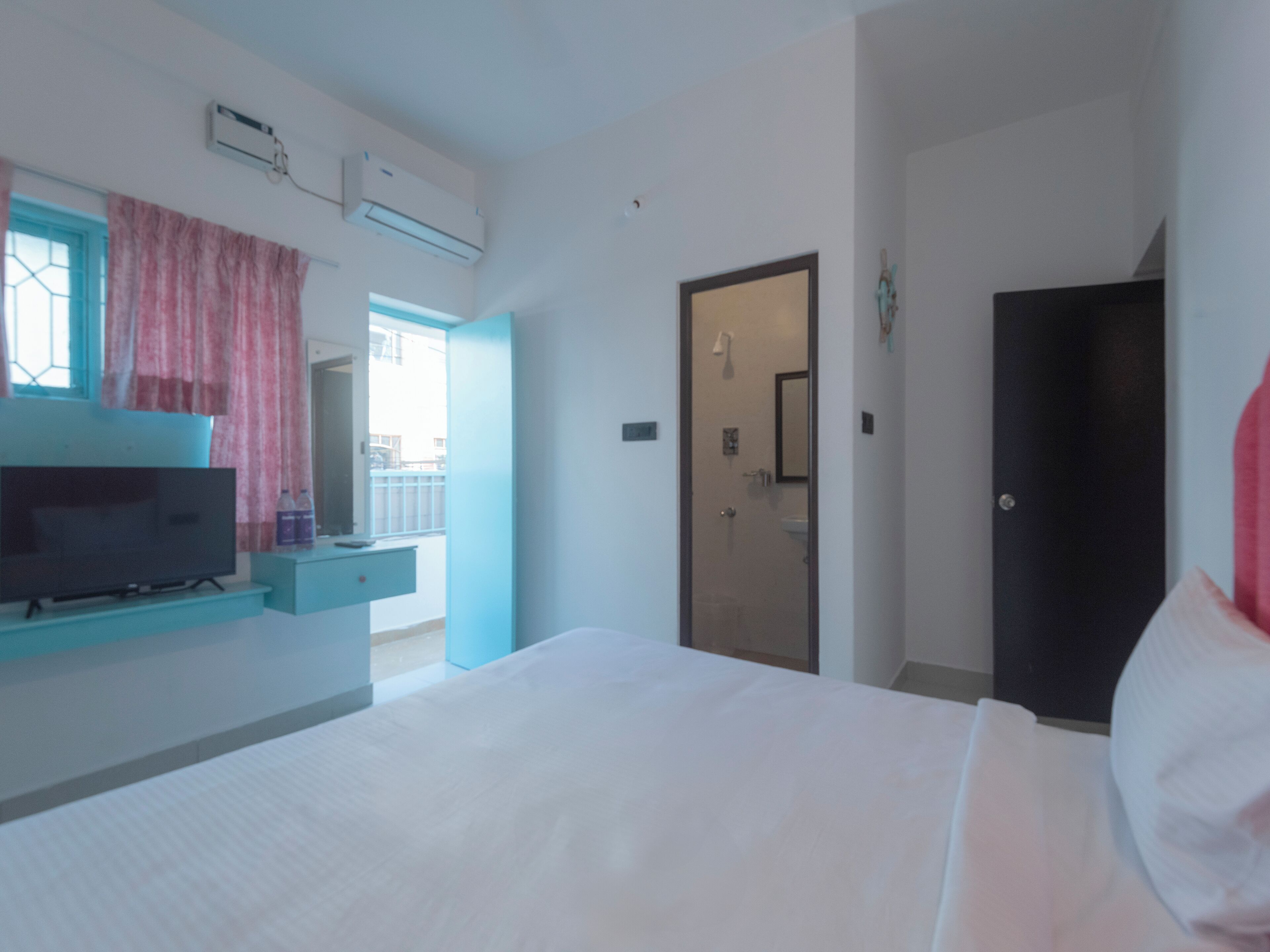 A hotel room with a bed, a TV, a window with curtains, an air conditioning unit, and a door leading to a bathroom.