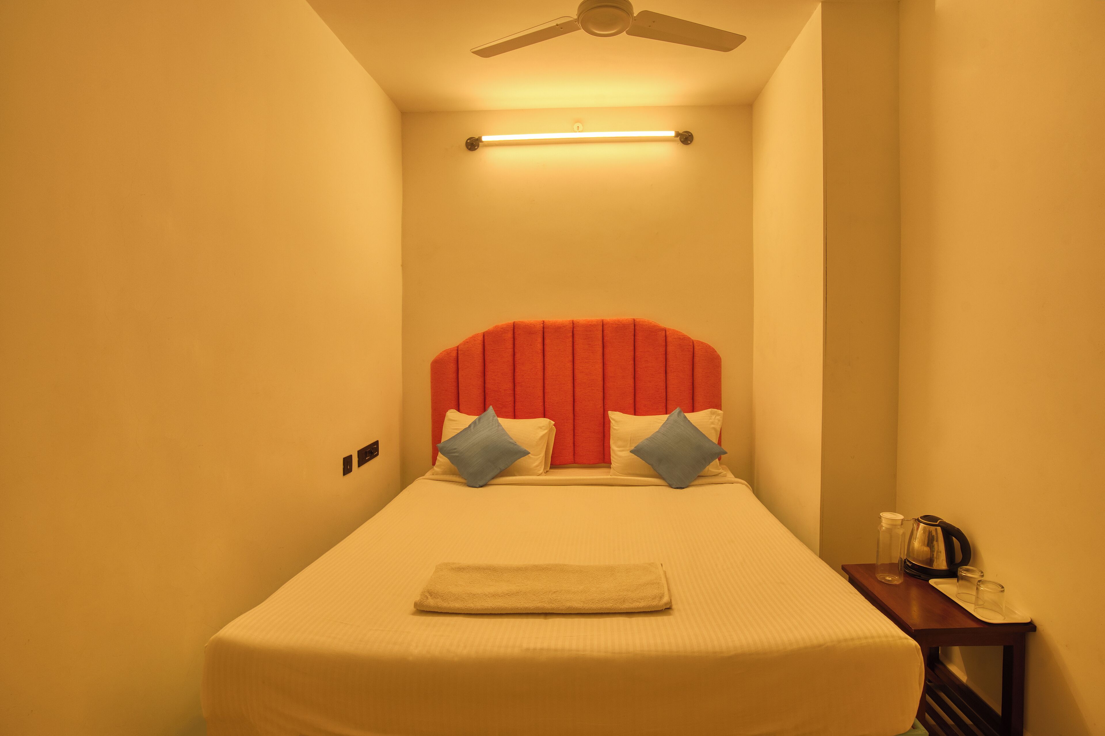 A small, single-bed room with a red headboard, two pillows, a bedside table with a kettle and glasses, and a ceiling fan.