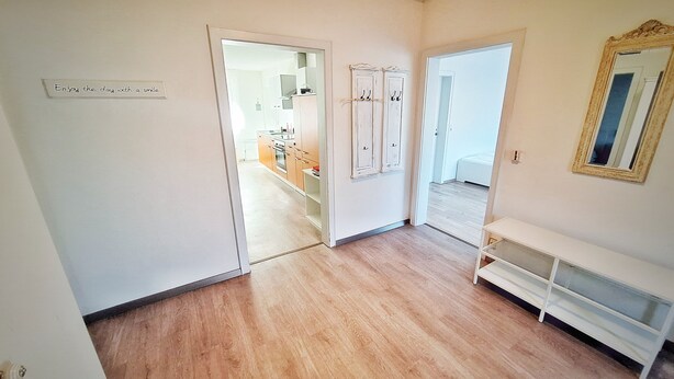 A hallway with wooden flooring, a white console table, and a mirror on the right. There is a door leading to a room with a bed and a bathroom visible through an open door. - Good prices Good travel, 4 bedrooms and 2 bathrooms on 130sqm (Neuried)