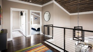Un interior moderno con techo de madera, una escalera con barandilla negra y vista a un dormitorio con cama y mesita de noche.