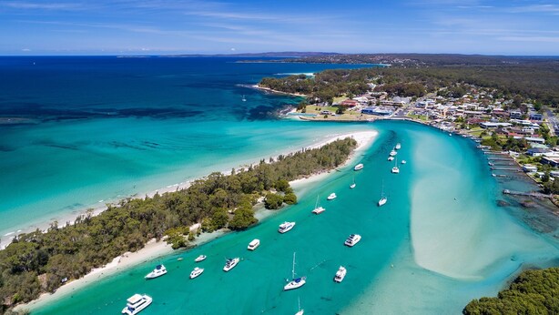 An aerial view of a coastal area with clear turquoise waters, a sandy beach, and numerous boats anchored near the shore.