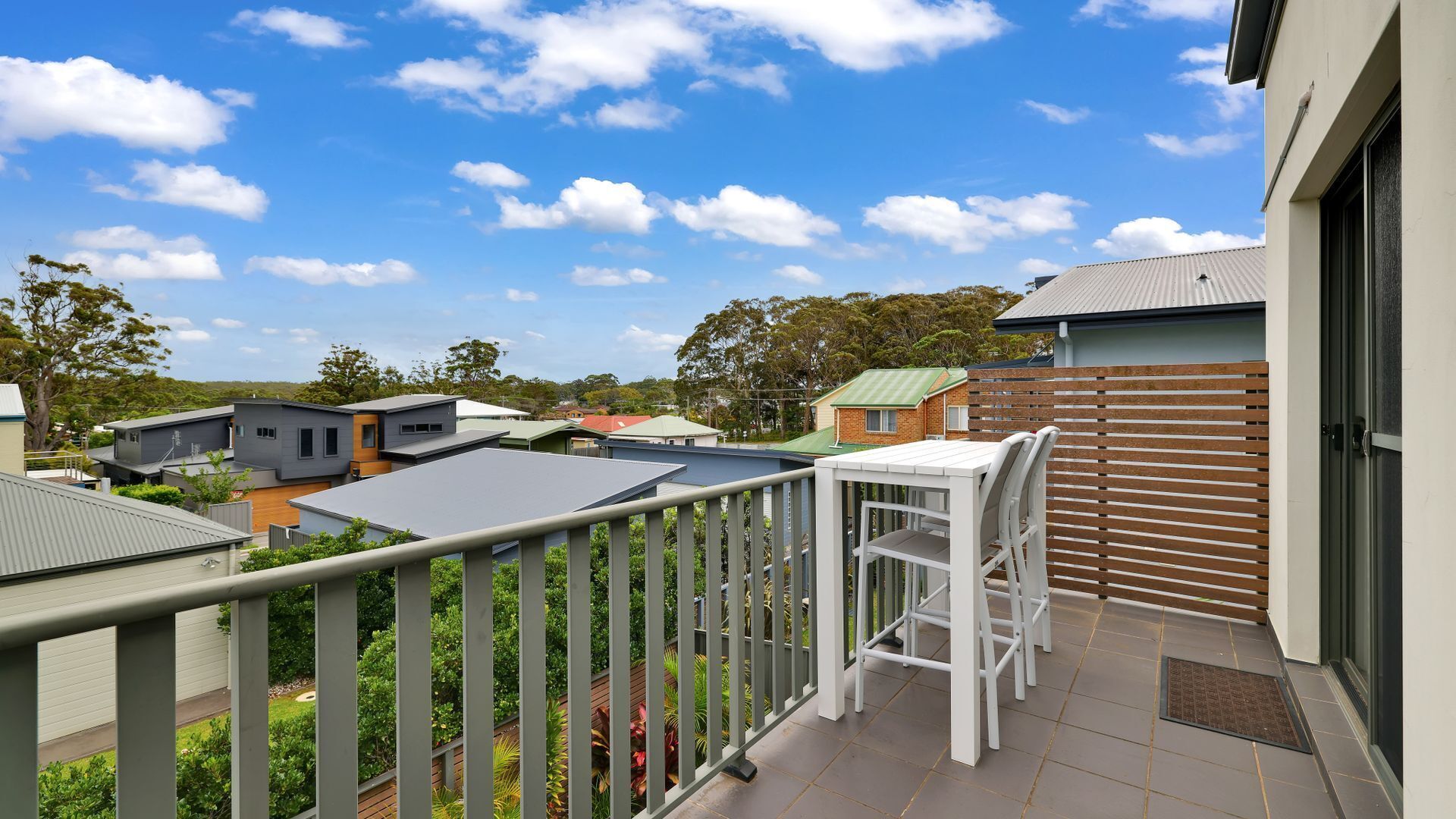 A balcony with a white table and chairs, a view of neighboring houses, and a clear blue sky.