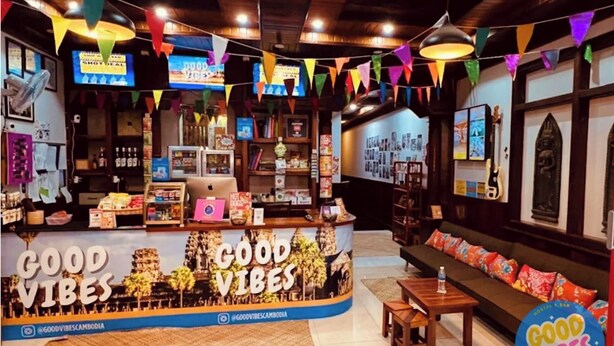 Un intérieur chaleureux et accueillant, avec un comptoir affichant le message « Bonne ambiance », des étagères remplies de livres et de souvenirs, un coin salon avec des coussins et un plafond en bois.