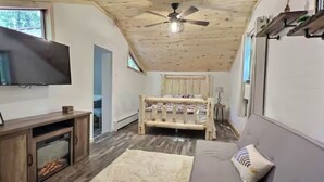 A cozy room with a wooden ceiling, a bed, a gray sofa, a TV, and a fireplace. - Cozy Cabin with Fireplace, Firepit, Close to Lake (Hawley)