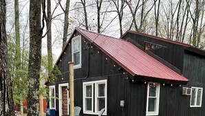 A black cabin with a red roof, a wooden deck, and a stone pathway. - Cozy Cabin with Fireplace, Firepit, Close to Lake (Hawley)