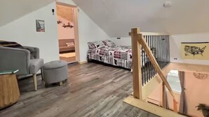 A loft bedroom with a sloped ceiling, a bed with a plaid bedspread, a gray sofa, and a wooden staircase. - Cozy Cabin with Fireplace, Firepit, Close to Lake (Hawley)
