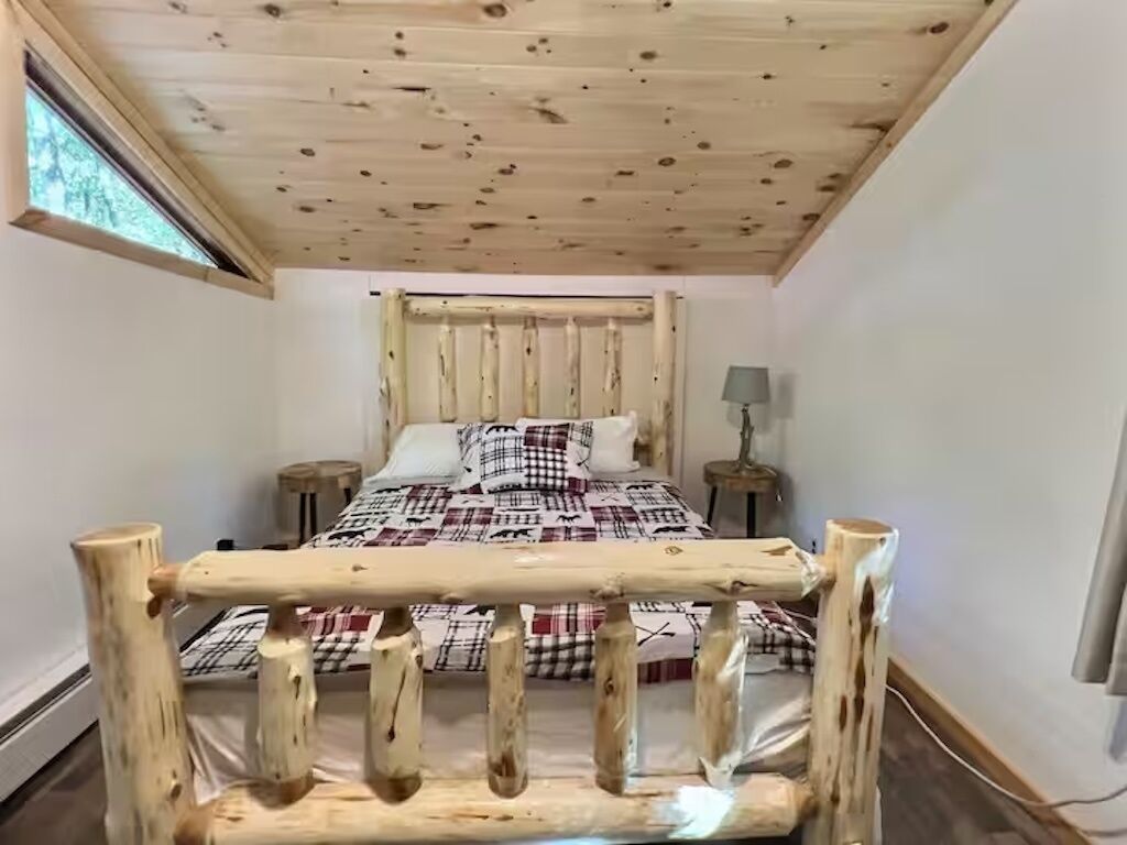 A bedroom with a wooden ceiling, a bed with a plaid bedspread, a nightstand, and a lamp.