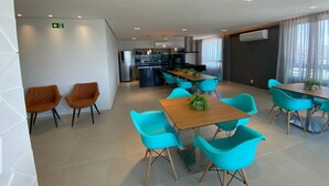 A modern dining area with a wooden table and turquoise chairs, a kitchen in the background, and a white wall with an emergency exit sign. - RN Studio Apto 414-Mandi Hospitalidade (Maceio)