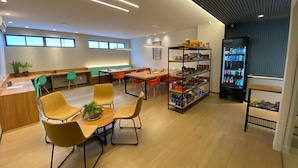 A modern lounge area with a wooden table, yellow chairs, a vending machine, and a bookshelf. - RN Studio Apto 414-Mandi Hospitalidade (Maceio)
