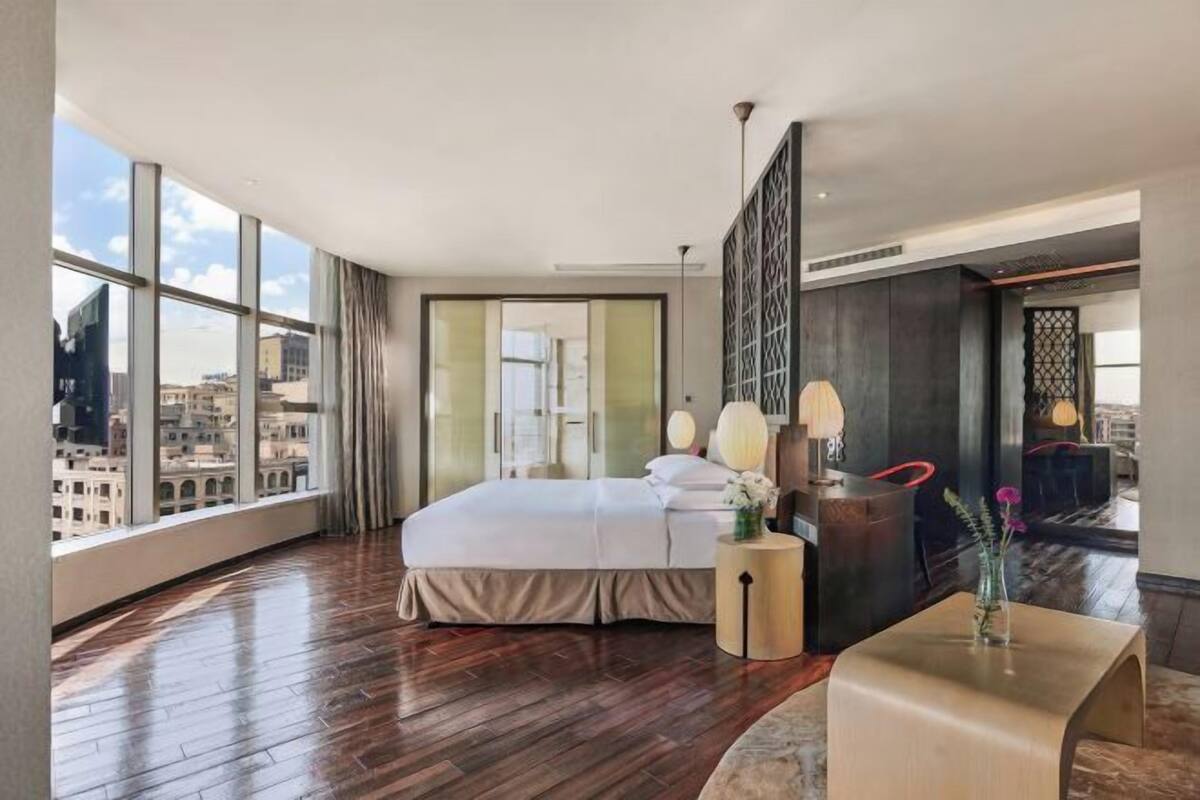 Een hotelkamer met een groot bed, een nachtkastje met lamp, uitzicht op de stad door grote ramen en een modern interieur.