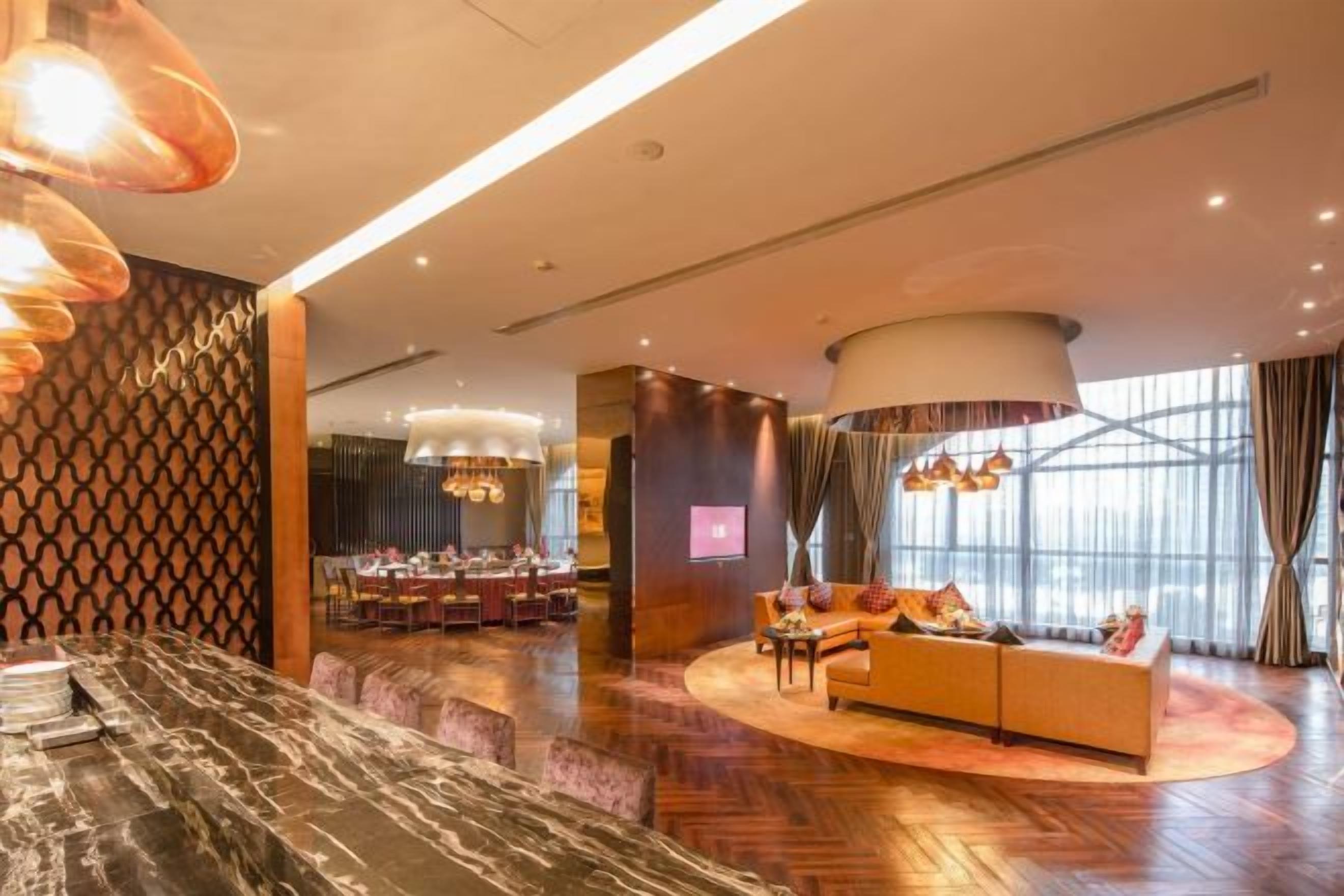 A modern hotel lobby with a marble reception desk, a dining area, a sofa, and a large window with curtains.